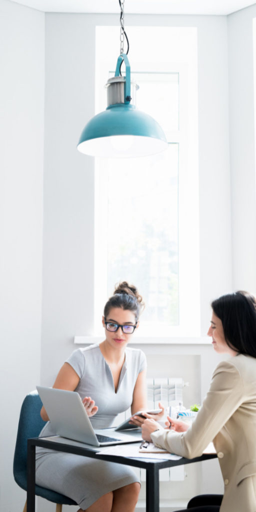 Two employees sitting in an office. Working at a small desk. One woman with glasses, and tied up hair points to the laptop screen. The second woman watches the screen.
