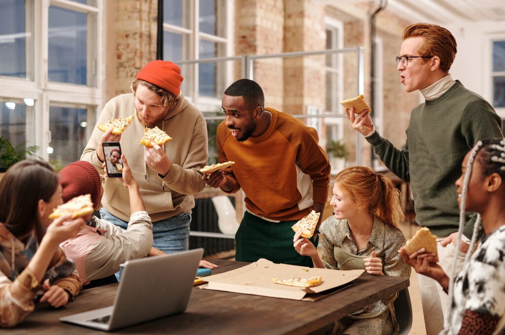 Group of happy employees smiling and eating pizza together in a bright, modern office, enjoying a casual team lunch